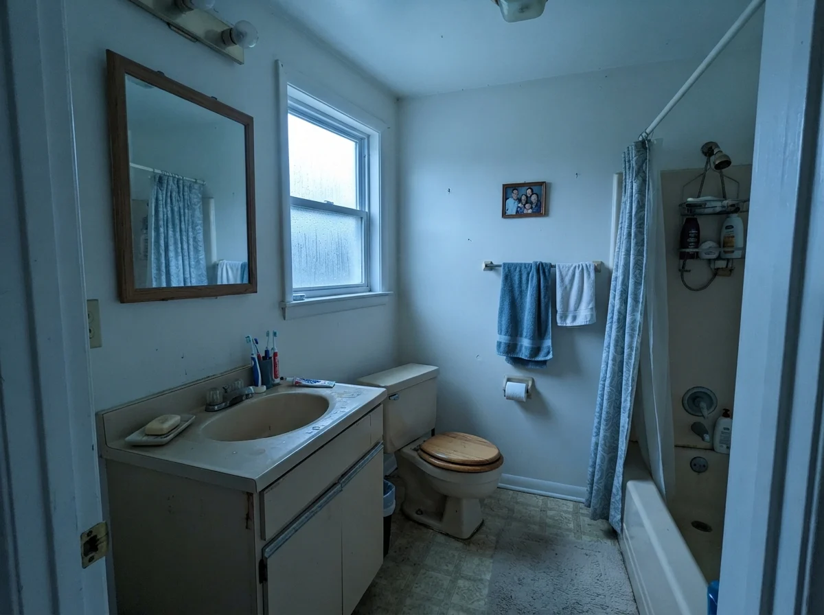 Modest dated bathroom with cool rainy north light casting on painted walls near an older beige vanity.